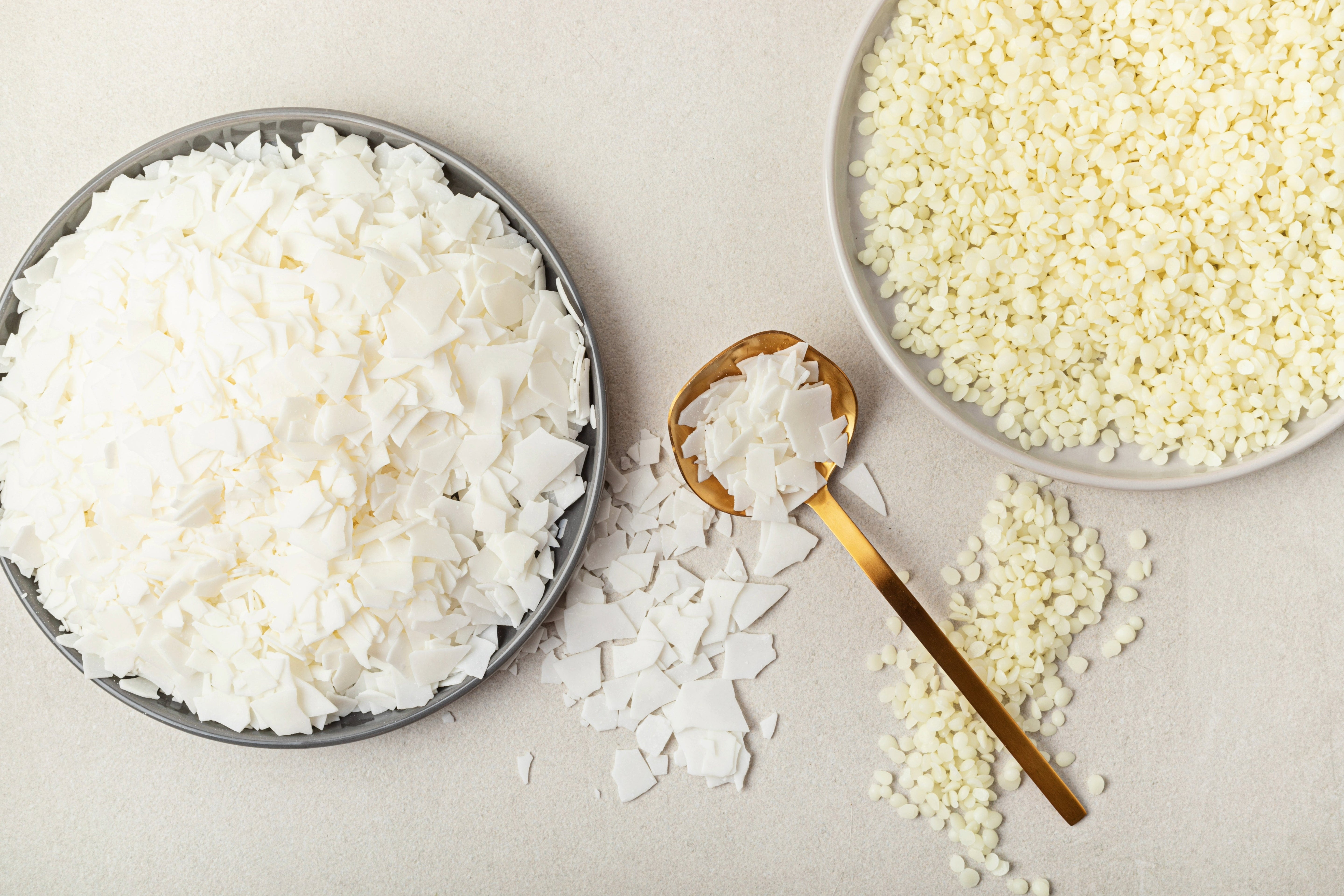 Verschiedene cremefarbene Flocken und Pastillen aus verschiedenen Wachsarten für Kerzen von oben fotografiert auf hellem Untergrund in verschiedenen Schalen