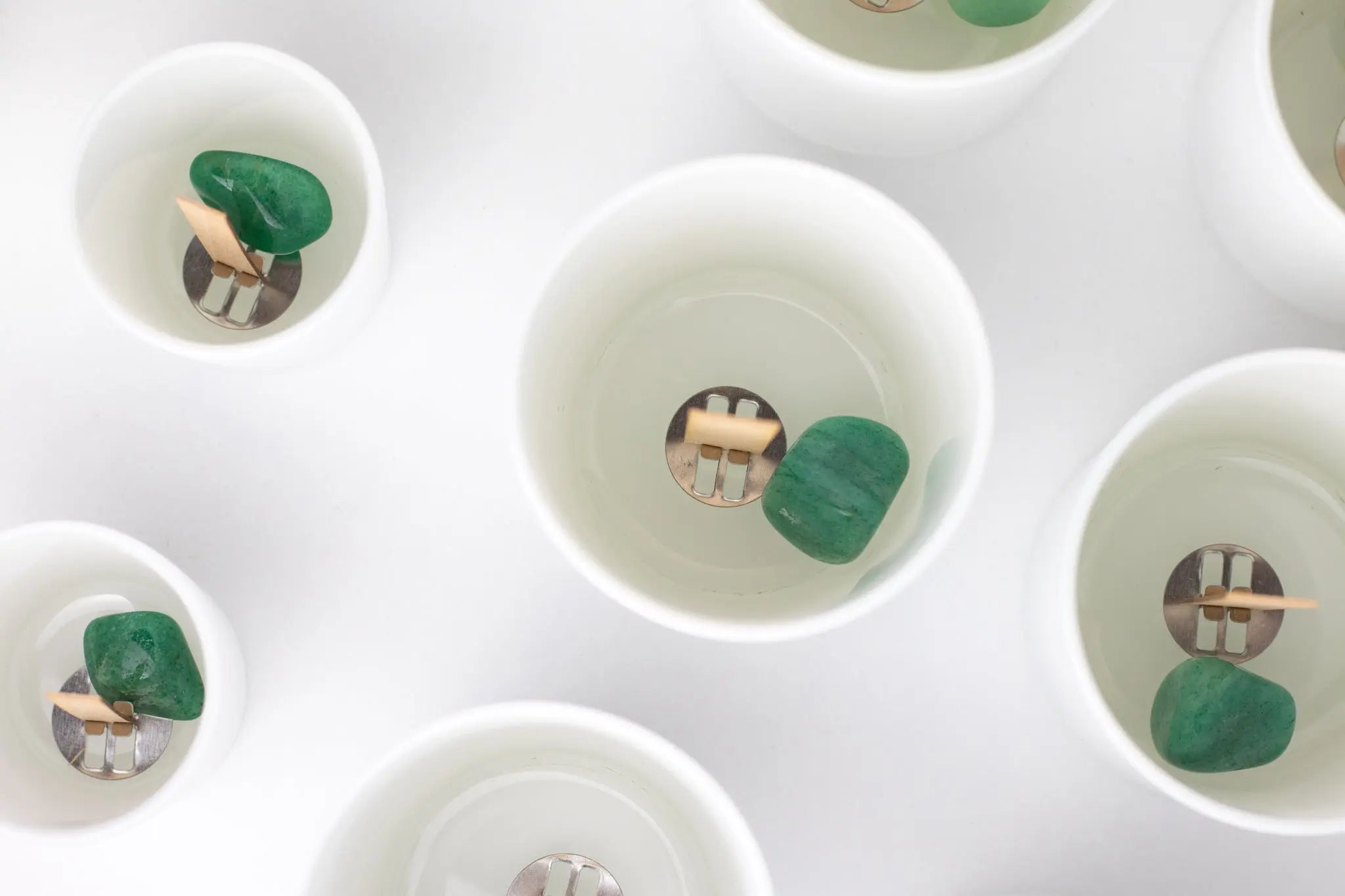 Weiße Kerzengläser vor der Herstellung von oben fotografiert, in denen sich mittig je ein Holzdocht mit Metall-Standfuß und daneben ein 25-45 mm großer Edelstein (Trommelstein eines grünen Aventurins) befindet